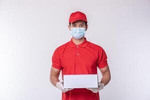 Immagine di un' consapevole giovane consegna uomo nel rosso berretto vuoto maglietta uniforme viso maschera guanti in piedi con vuoto bianca cartone scatola isolato su leggero grigio sfondo studio foto