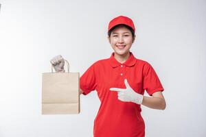 Immagine di un' contento giovane consegna uomo nel rosso berretto vuoto maglietta uniforme in piedi con vuoto Marrone mestiere carta pacchetto isolato su leggero grigio sfondo studio foto