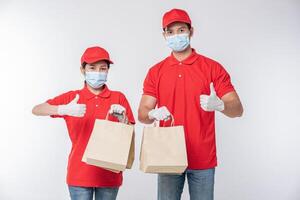 Immagine di un' contento giovane consegna uomo nel rosso berretto vuoto maglietta uniforme viso maschera guanti in piedi con vuoto Marrone mestiere carta pacchetto isolato su leggero grigio sfondo studio foto