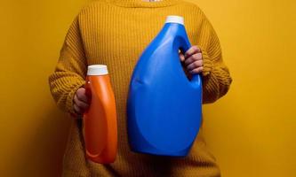donna nel un' a maglia maglione detiene un arancia e blu plastica bottiglie con liquido lavaggio gel. lavanderia e lavoretti, giallo sfondo foto