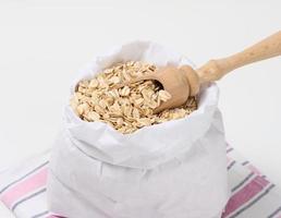 crudo fiocchi d'avena nel un' bianca carta Borsa e un' di legno cucchiaio su un' bianca tavolo, prima colazione porridge foto