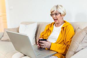 ritratto di anziano donna seduta a casa su il divano e Lavorando su sua il computer portatile. più vecchio signora fare surf il netto o avendo video chiamata a partire dal casa e sensazione eccitante. Tenere tazza di caffè o tè foto