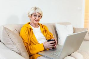 anziano donna spende casa tempo libero con un' il computer portatile. smilling anziano donna si siede a un' accogliente divano, Tenere tazza di caffè o tè. ragnatela navigando, Guardando tv serie, shopping nel rete,video chiamata foto