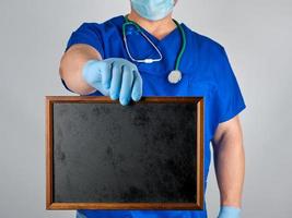 medico nel blu uniforme e sterile latice guanti Tenere un' di legno telaio con un' vuoto nero lettering sfondo foto