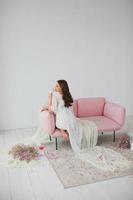 giovane sposa nel bianca vestaglia in posa su il rosa divano nel il studio foto