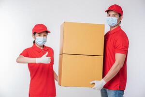 Immagine di un' consapevole giovane consegna uomo nel rosso berretto vuoto maglietta uniforme viso maschera guanti in piedi con vuoto Marrone cartone scatola isolato su leggero grigio sfondo studio foto