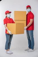 Immagine di un' consapevole giovane consegna uomo nel rosso berretto vuoto maglietta uniforme viso maschera guanti in piedi con vuoto Marrone cartone scatola isolato su leggero grigio sfondo studio foto