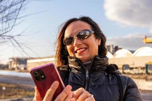 mezzo anziano donna indossare inverno Abiti utilizzando smartphone nel davanti di un' fabbrica foto