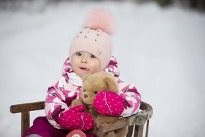 un' poco ragazza si siede nel un' di legno slitta con un' orsacchiotto orso nel inverno. foto