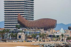Franco Gehry's d'oro pesce scultura e edifici nel davanti di sabbioso e roccioso spiaggia con mapfre Torre nel il sfondo a porta olimpico nel catalogna, Barcellona, Spagna foto