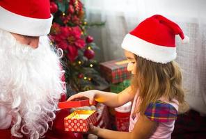 poco ragazza nel Santa cappello su Babbo Natale giro vicino Natale albero nel Natale arredamento. Spettacoli un' scatola con un' regalo, un' morbido giocattolo, tocchi il suo barba e ride. nuovo anno, russo nonno brina foto