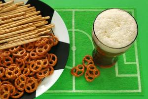 calcio calcio festa tavolo con spuntini e bevanda. salato salatino e cannucce su grande piatto dipinto piace calcio sfera, bicchiere di birra, su piatto stuoia piace calcio campo fatto di verde provato, superiore Visualizza. foto