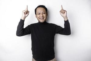 eccitato asiatico uomo indossare nero camicia puntamento a il copia spazio su superiore di lui, isolato di bianca sfondo foto