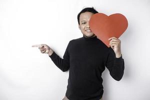 eccitato asiatico uomo indossare nero camicia, puntamento a il copia spazio accanto lui mentre Tenere un' grande rosso a forma di cuore carta, isolato di bianca sfondo foto