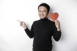 eccitato asiatico uomo indossare nero camicia, puntamento a il copia spazio accanto lui mentre Tenere un' grande rosso a forma di cuore carta, isolato di bianca sfondo foto