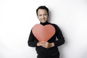 un' ritratto di un' contento asiatico uomo indossare un' nero camicia, Tenere un' rosso a forma di cuore carta isolato di bianca sfondo foto