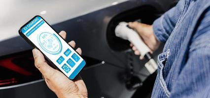 uomo ricarica elettrico veicolo con cavo guardare a App su mobile Telefono foto