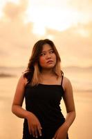 un asiatico donna pose con un' sporco e arrabbiato espressione quando indossare un' nero vestito su il spiaggia foto