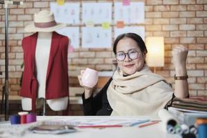 asiatico maturo femmina moda progettista chi sorrisi e sembra a telecamera celebra e è allegro con vestito design opera successo e gioioso nel stilista studio, un' contento costume boutique piccolo attività commerciale. foto