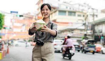 contento giovane asiatico donna zaino viaggiatore potabile un' Noce di cocco succo a Cina cittadina strada cibo mercato nel bangkok, Tailandia. viaggiatore controllo su lato strade. foto