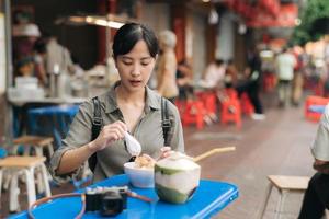 contento giovane asiatico donna zaino viaggiatore godendo strada cibo a Cina cittadina strada cibo mercato nel bangkok, Tailandia. viaggiatore controllo su lato strade. foto