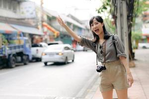 sorridente giovane asiatico donna viaggiatore autostop su un' strada nel il città. vita è un' viaggio concetto. foto