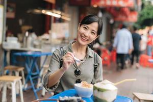 contento giovane asiatico donna zaino viaggiatore godendo strada cibo a Cina cittadina strada cibo mercato nel bangkok, Tailandia. viaggiatore controllo su lato strade. foto