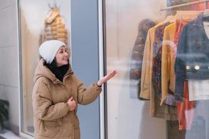 un' elegante giovane donna nel un' cappello e cappotto nel il città sembra in il finestre di capi di abbigliamento I negozi foto