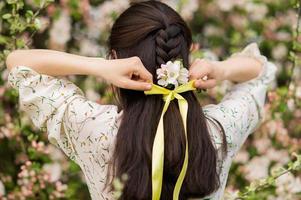 un' giovane donna cravatte un' arco su sua capelli con un' giallo nastro nel un' fioritura giardino foto