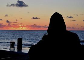 maschio silhouette nel un' cappuccio a un' tavolo nel un' bar contro il fondale di il tramonto mare foto