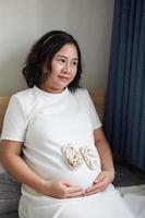 foto di contento asiatico incinta donna seduta nel letto e toccante sua pancia a casa con piccolo scarpe per il nascituro bambino nel il pancia