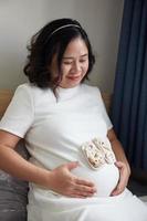 foto di contento asiatico incinta donna seduta nel letto e toccante sua pancia a casa . con piccolo scarpe per il nascituro bambino nel il pancia