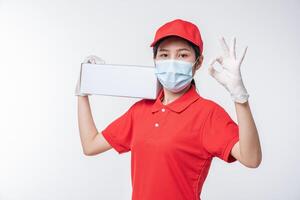 Immagine di un' consapevole giovane consegna uomo nel rosso berretto vuoto maglietta uniforme viso maschera guanti in piedi con vuoto bianca cartone scatola isolato su leggero grigio sfondo studio foto