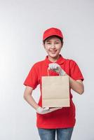 Immagine di un' contento giovane consegna uomo nel rosso berretto vuoto maglietta uniforme in piedi con vuoto Marrone mestiere carta pacchetto isolato su leggero grigio sfondo studio foto