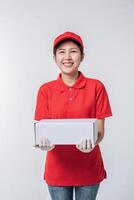 Immagine di giovane consegna uomo nel rosso berretto vuoto maglietta uniforme in piedi con vuoto bianca cartone scatola isolato su leggero grigio sfondo studio foto