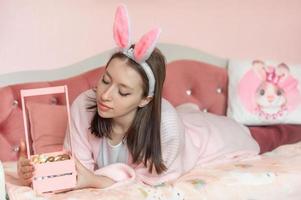 un' bellissimo giovane ragazza con lepre orecchie su sua testa bugie su il letto a casa con un' cestino di decorato Pasqua uova. preparazione per est foto