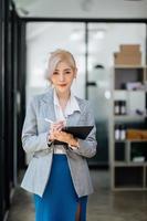 fiducioso asiatico donna con un' Sorridi in piedi Tenere bloc notes e tavoletta a il ufficio. foto
