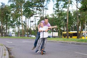 un' padre cavalcate il suo figlio nel un' bianca maglietta su un' scooter, il contento bambino diffusione il suo mani foto