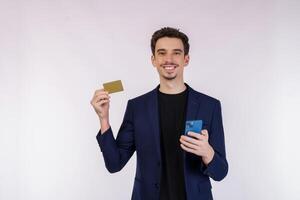 ritratto di contento giovane uomo d'affari in piedi utilizzando mobile cellula Telefono e Tenere credito banca carta isolato su bianca colore sfondo studio foto
