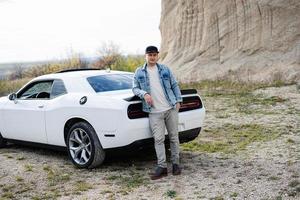 bello uomo nel jeans giacca e berretto è in piedi vicino il suo bianca muscolo auto nel carriera. foto