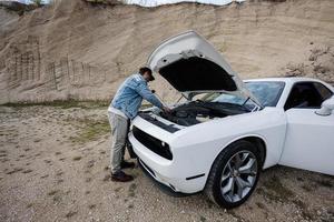 bello uomo nel jeans giacca e berretto è in piedi vicino il suo bianca muscolo auto con apertura cappuccio, dai un'occhiata olio livello di motore. foto