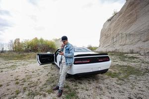 bello uomo nel jeans giacca e berretto, con mobile Telefono è in piedi vicino il suo bianca muscolo auto nel carriera. foto