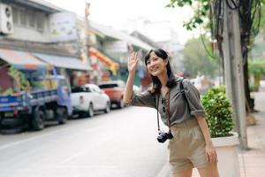 sorridente giovane asiatico donna viaggiatore autostop su un' strada nel il città. vita è un' viaggio concetto. foto