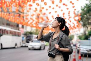 contento giovane asiatico donna zaino viaggiatore potabile un' freddo acqua a Cina cittadina strada cibo mercato nel bangkok, Tailandia. viaggiatore controllo su lato strade. foto