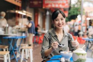 contento giovane asiatico donna zaino viaggiatore godendo strada cibo a Cina cittadina strada cibo mercato nel bangkok, Tailandia. viaggiatore controllo su lato strade. foto