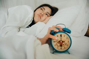 asiatico bellissimo donna nel bianca pigiama svolta via allarme orologio mentre assonnato nel letto a sua Camera da letto di Casa su vacanze. ragazza è avendo guaio scia su in ritardo nel mattina. malsano dormire. selettivo messa a fuoco foto