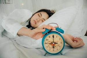 asiatico bellissimo donna nel bianca pigiama svolta via allarme orologio mentre assonnato nel letto a sua Camera da letto di Casa su vacanze. ragazza è avendo guaio scia su in ritardo nel mattina. malsano dormire. selettivo messa a fuoco foto