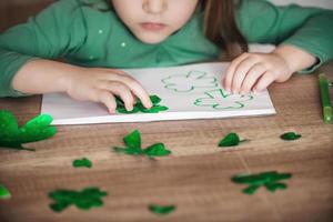 un' bambino a casa a il tavolo disegna e tagli verde quadrifogli per st. Patrick giorno. foto