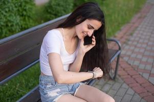 il ragazza è seduta su un' panca, ridendo e guardare a il Telefono, Là è un' il computer portatile nelle vicinanze foto