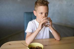 un' carino ragazzo bevande acqua e mangia un' asciutto prima colazione a casa e sorrisi foto